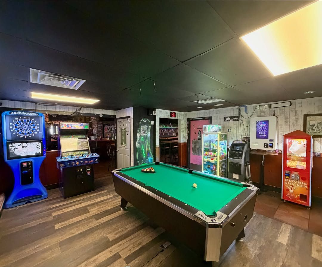 Green felt pool table and various arcade games in a dimly lit game room.