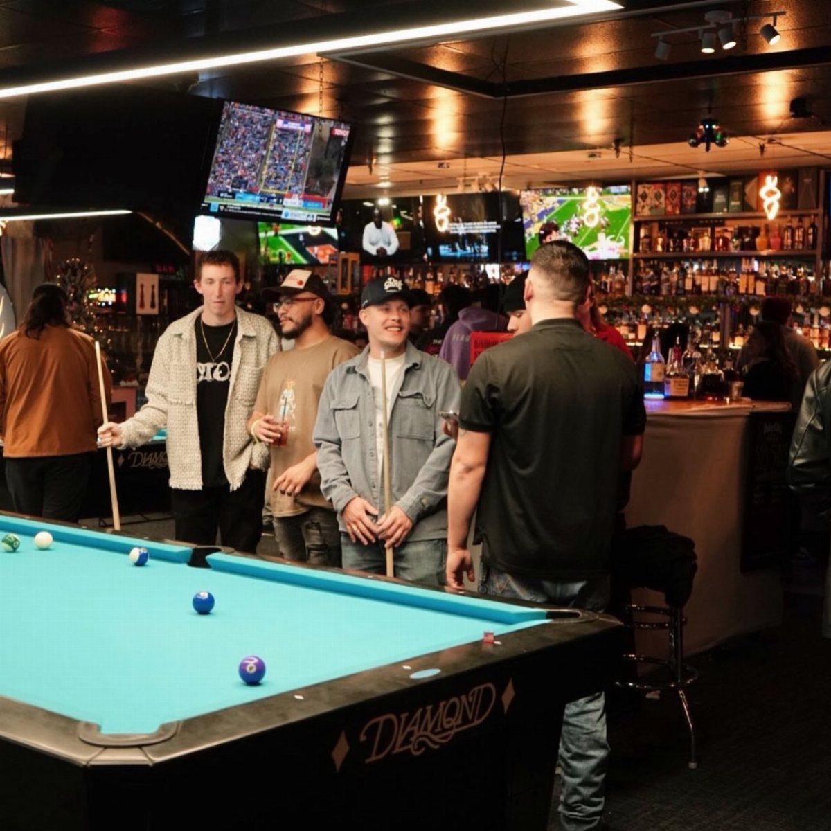 Men standing by a blue pool table in a bar with sports on TV.