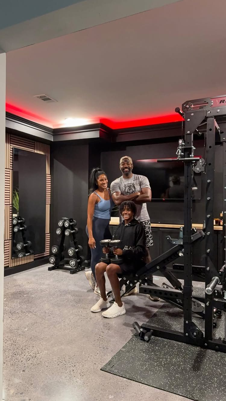 Three people posing in a modern basement gym with weightlifting equipment and red lighting.