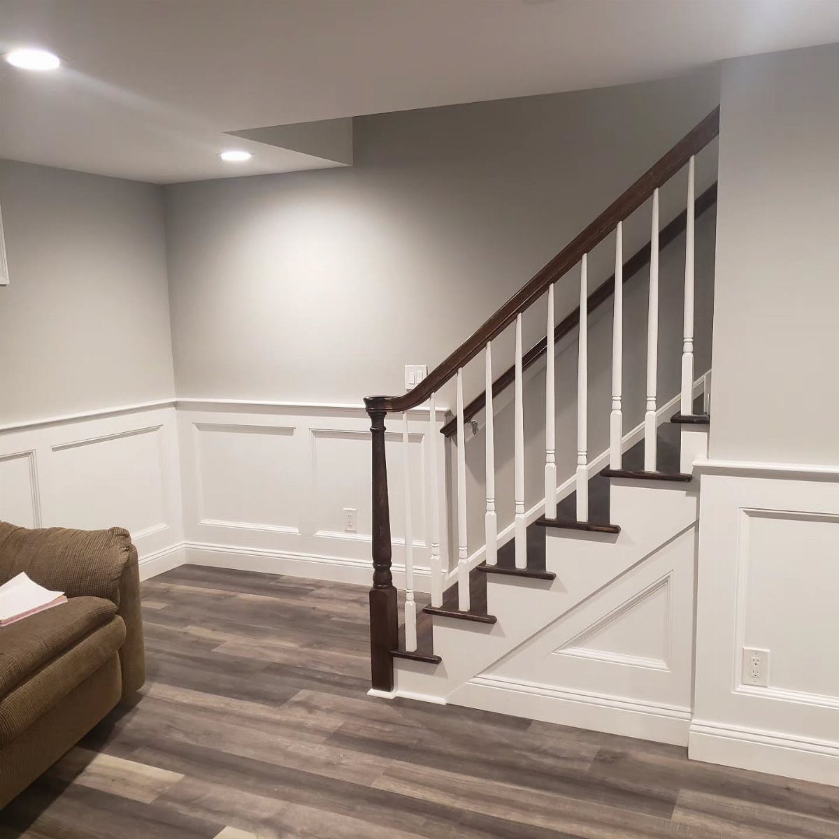 Finished basement with grey walls, white wainscoting, and a dark wood staircase railing.