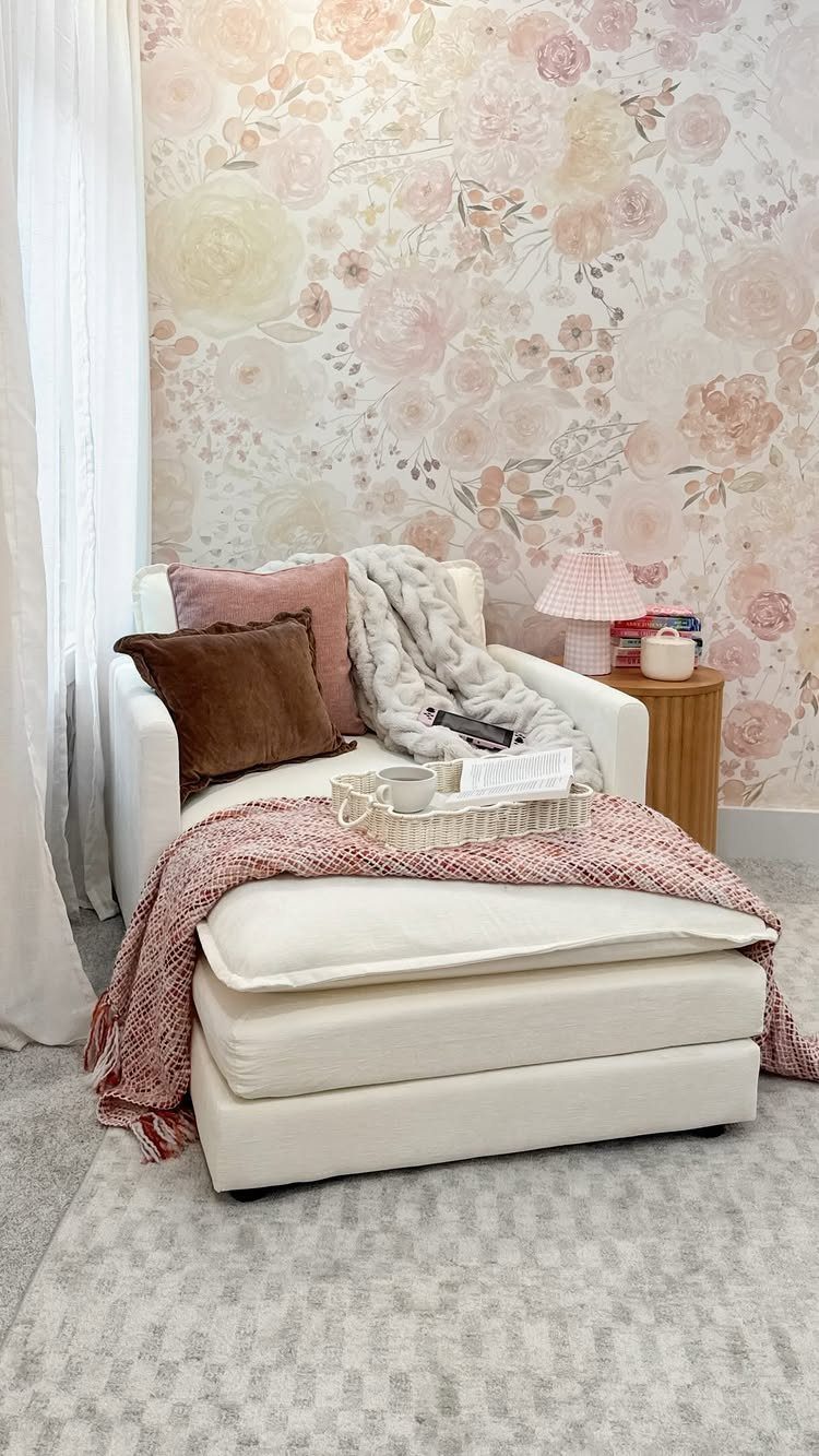 White chaise lounge with plush pillows and blankets against floral wallpaper.