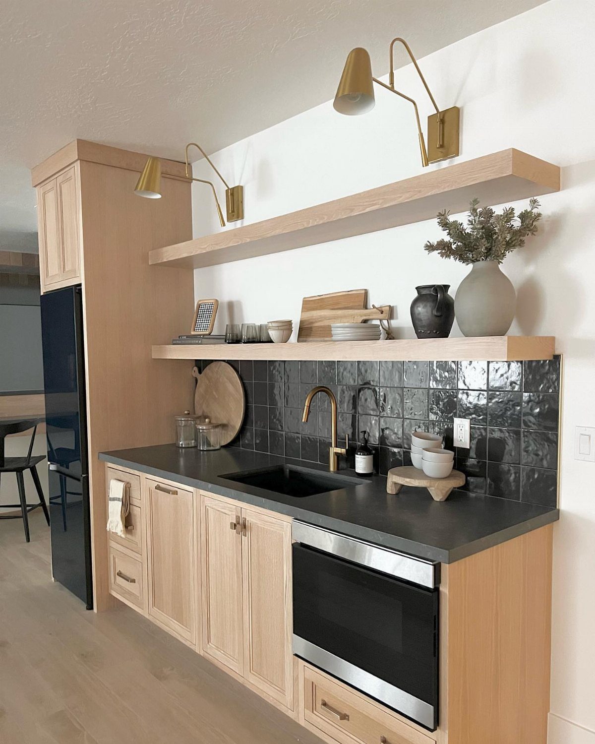 Compact kitchenette bar with light wood cabinets, dark countertop, and gold wall lighting.