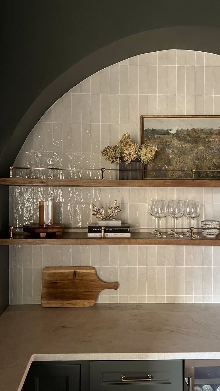 Light vertical tile wall behind wooden floating shelves with glassware and a painting.
