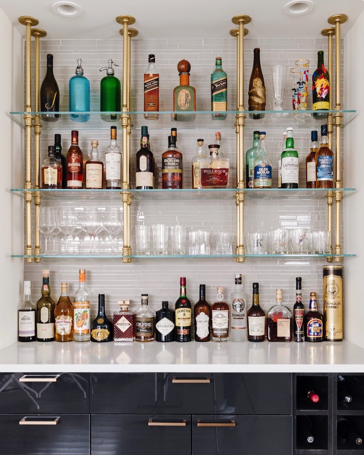 Glass shelves with gold supports holding liquor bottles and glassware against white tile.
