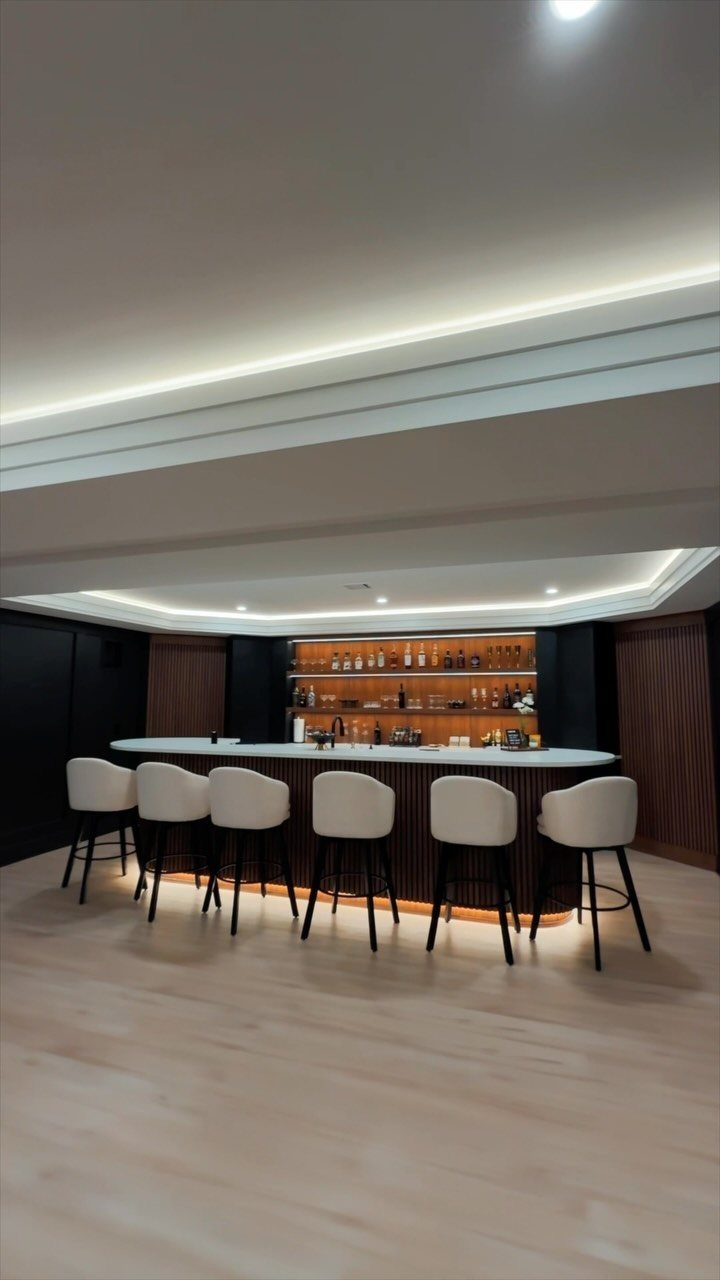 Curved home bar with white stools and backlit shelves in a modern basement.