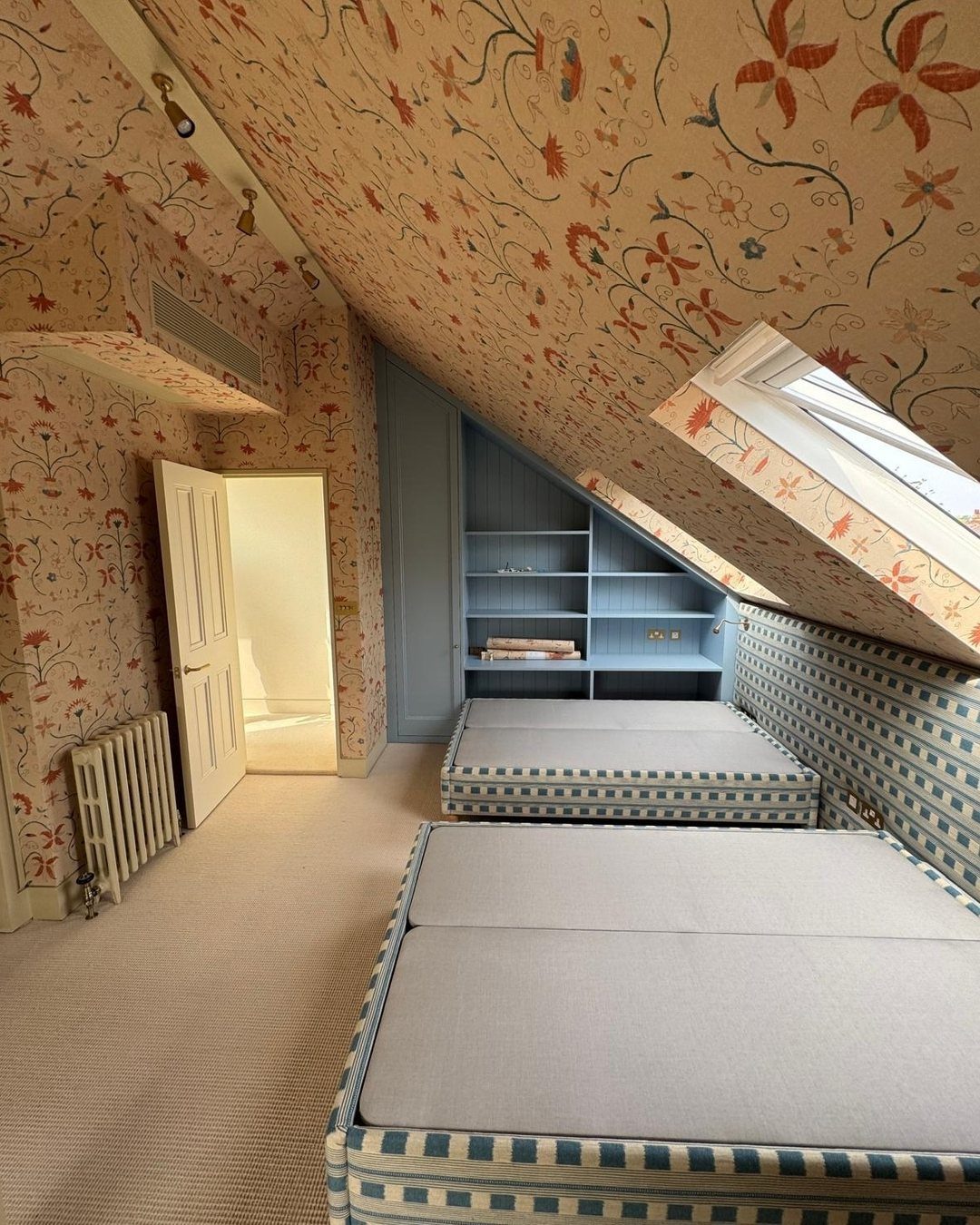 Attic bedroom with floral wallpaper and twin beds with blue checkered textile bases.