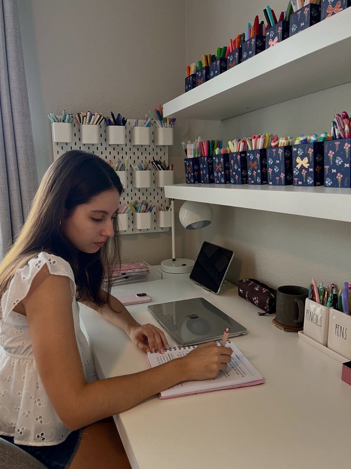 Young woman writing in a notebook at a white desk with organized markers.