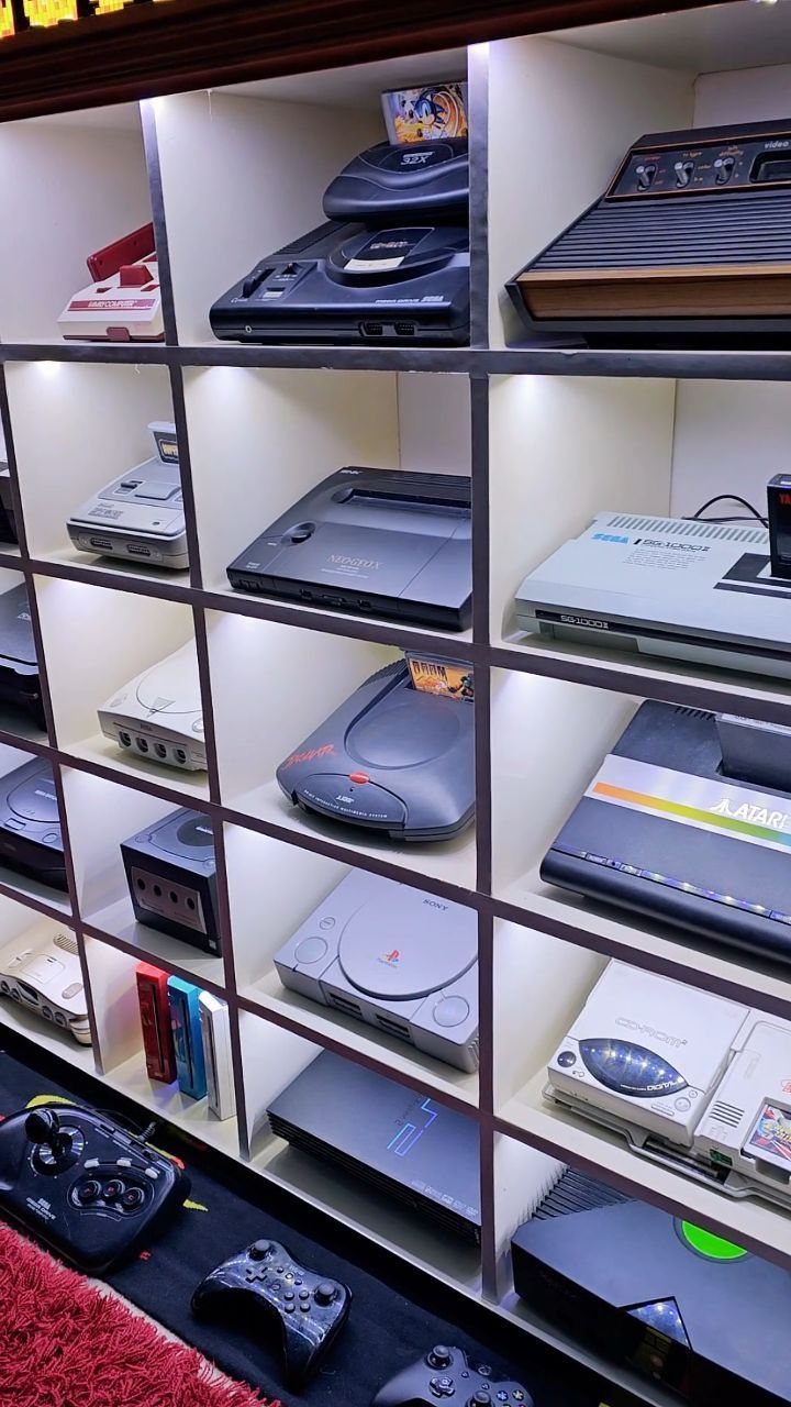 Collection of vintage gaming consoles displayed on illuminated white shelving with controllers below.