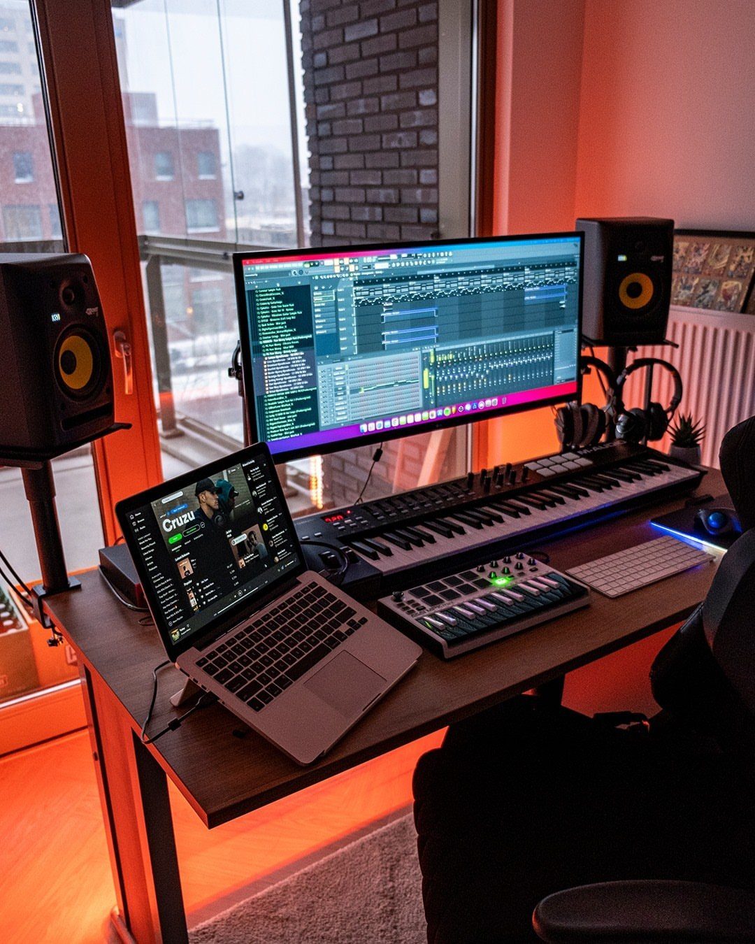 Music production setup with a large monitor and laptop on a wooden desk.