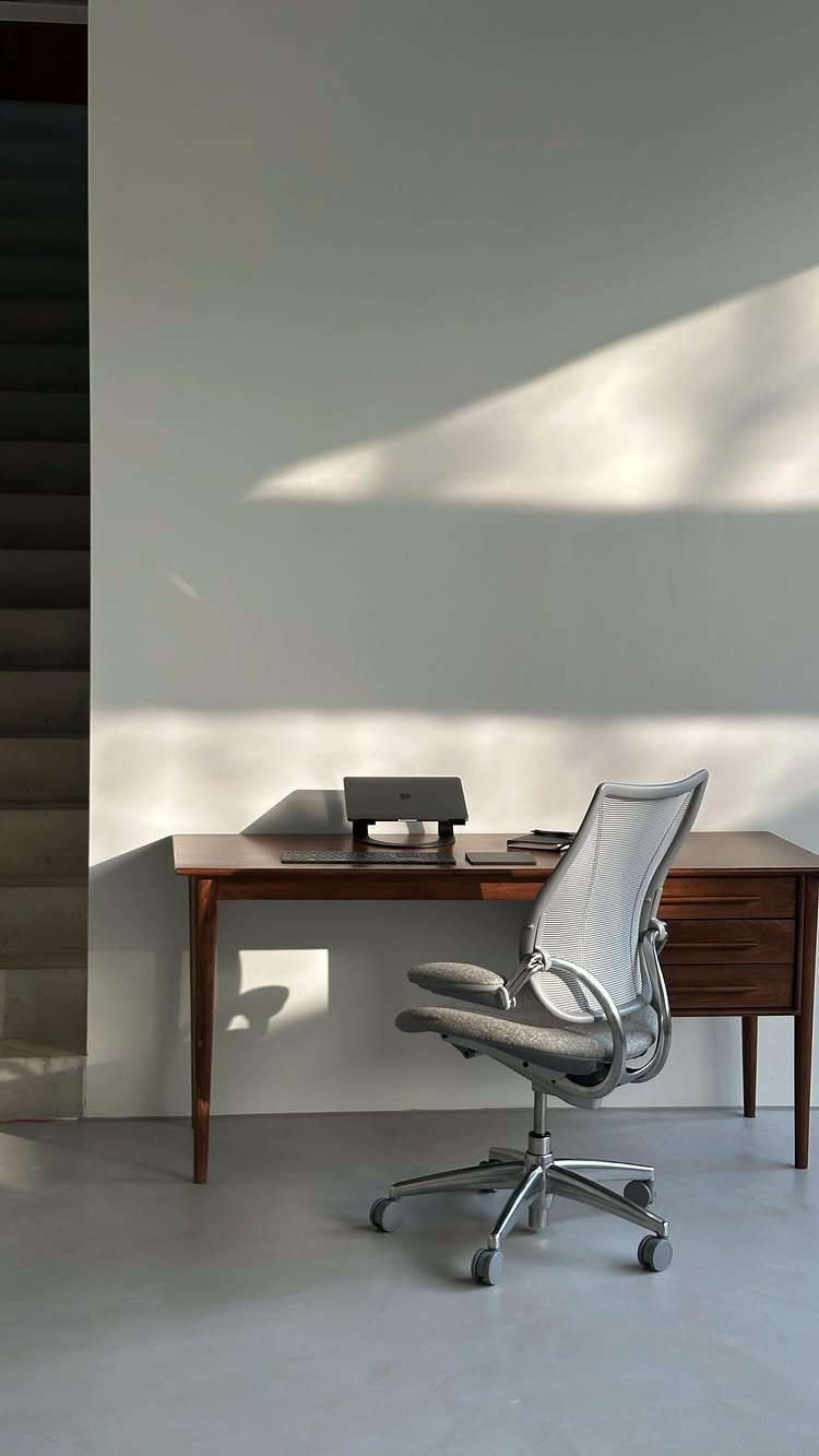 White mesh ergonomic office chair next to a dark wood desk in a bright room.