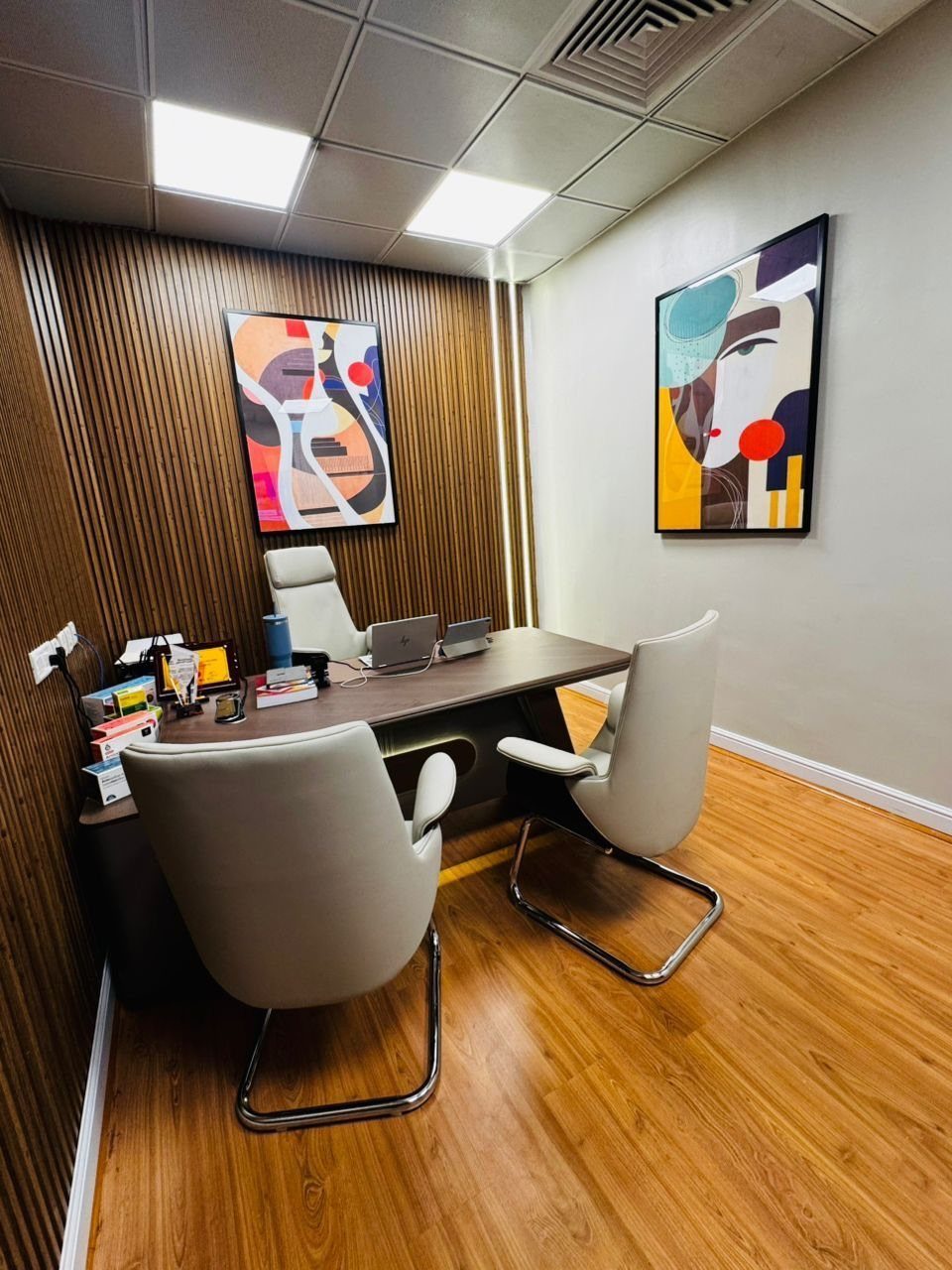 Modern home office with a wooden desk, white chairs, and abstract wall art.