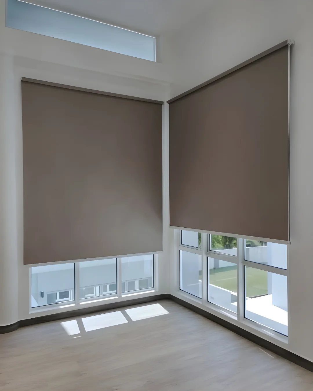 Taupe roller blinds partially covering large white-framed windows in a bright empty room.