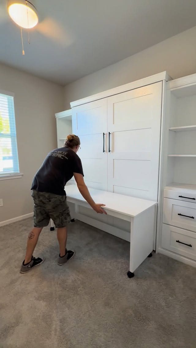 Man standing by a white built-in murphy bed and desk unit in a room.