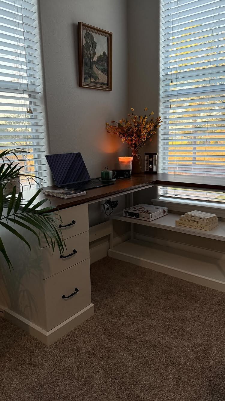 Built-in L-shaped corner desk with dark wood top and white drawers between two windows.
