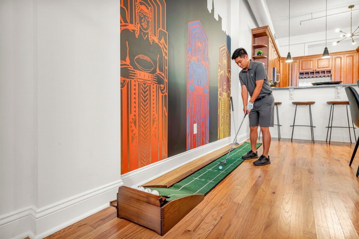 Man playing indoor mini golf on a green turf track beside a colorful mural.