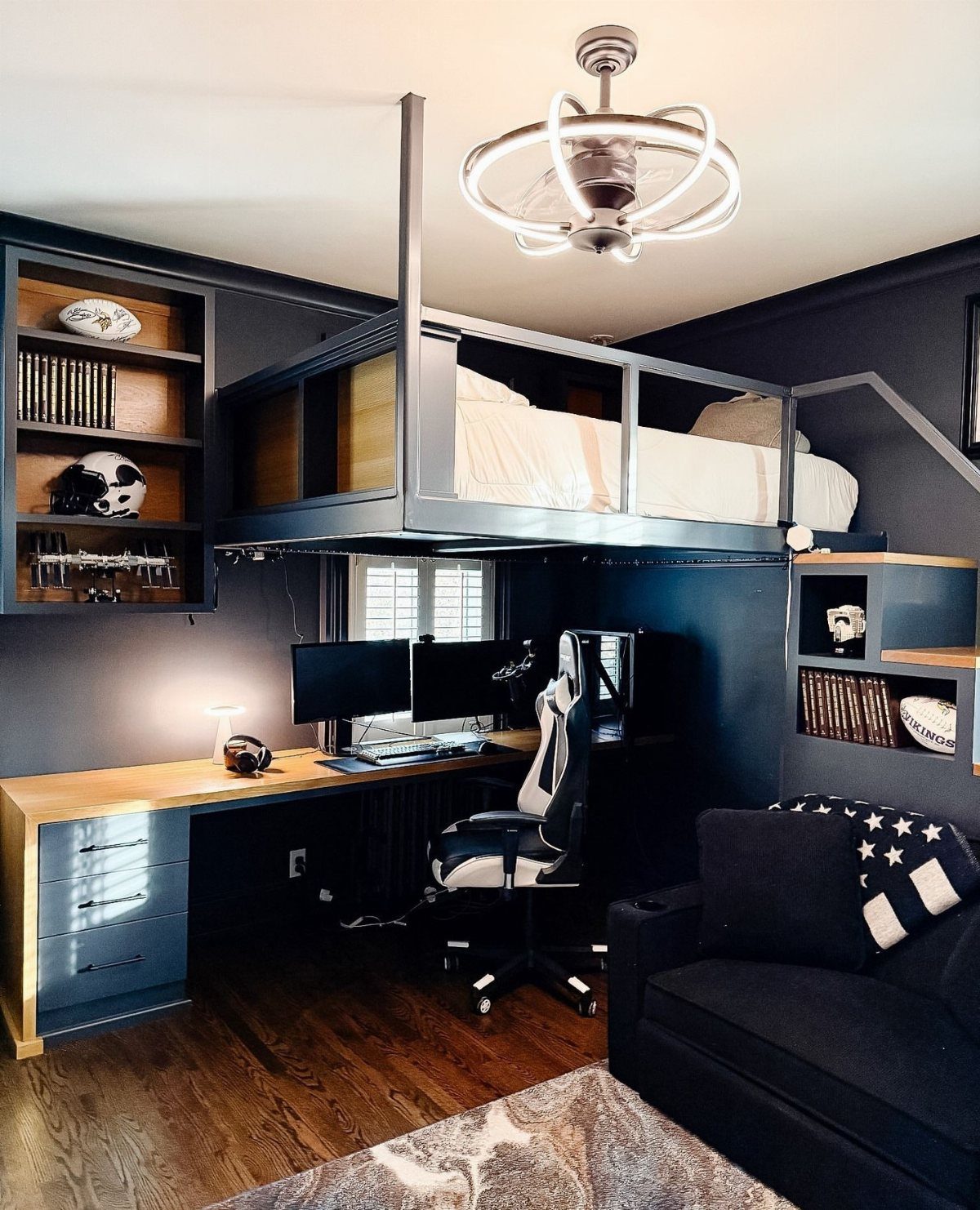 Gaming setup with dual monitors under a loft bed in a dark grey room.