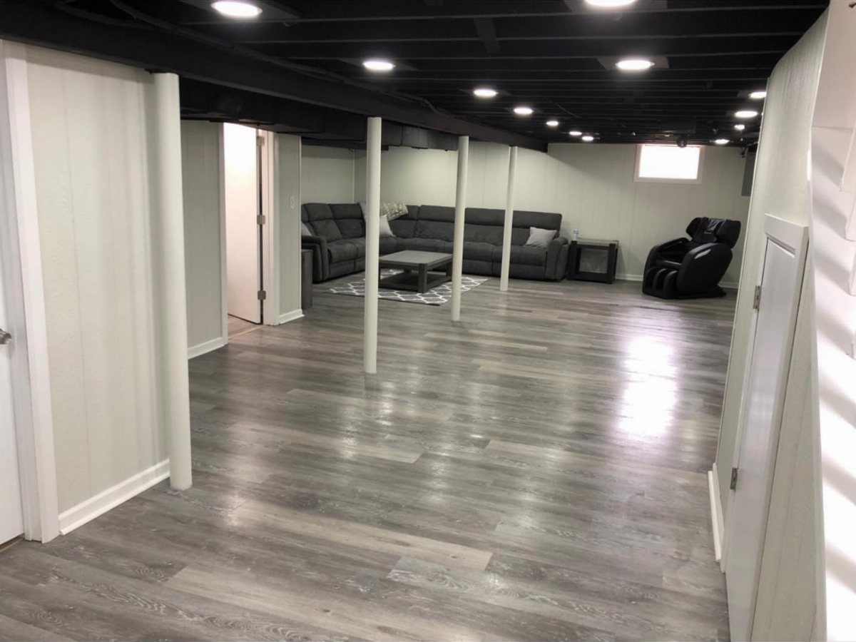 Finished basement with black exposed ceiling joists, grey flooring, and a grey sectional sofa.