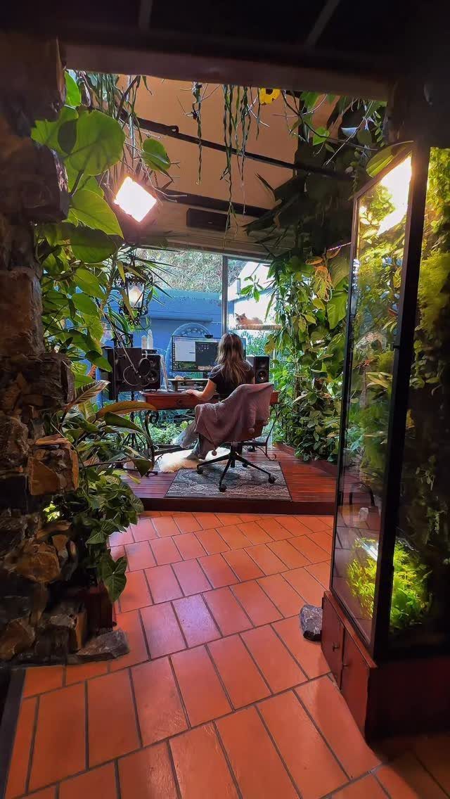 Home office workspace surrounded by dense green plants and a large vertical terrarium.