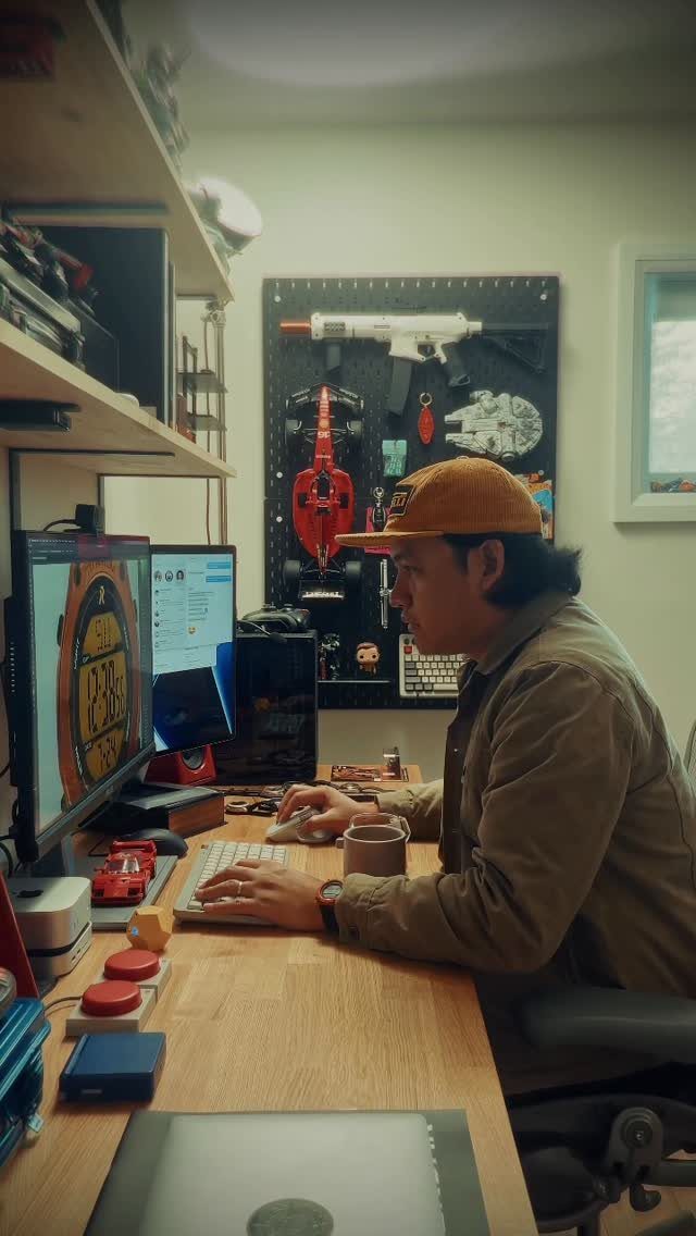Man sitting at a wooden desk with dual monitors and a tool pegboard.