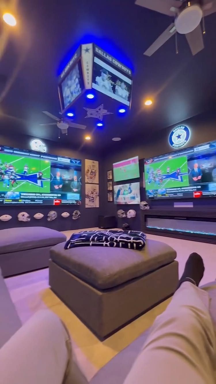 Gaming room with multiple screens, blue LED ceiling lights, and grey ottomans.