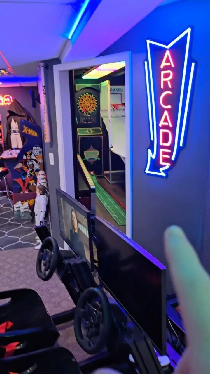 Gaming room with blue neon arcade sign, LED ceiling lights, and racing simulator setup.