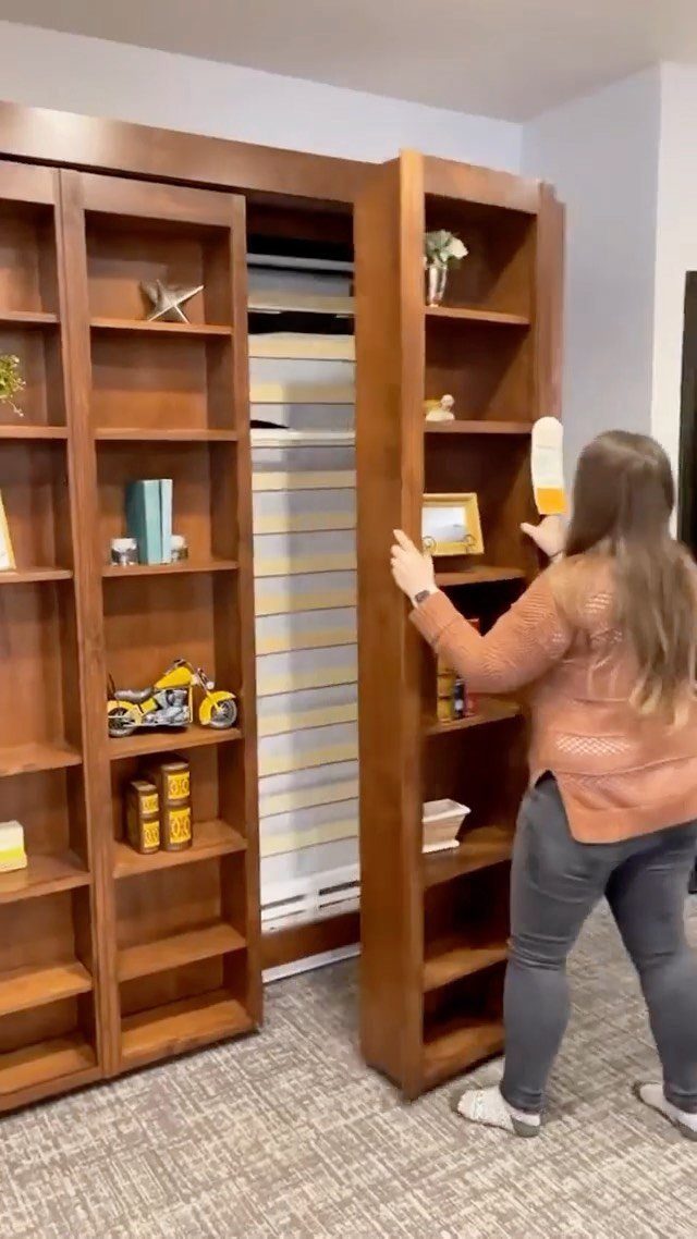 Woman opening a wooden bookshelf to reveal a hidden murphy bed in a room.