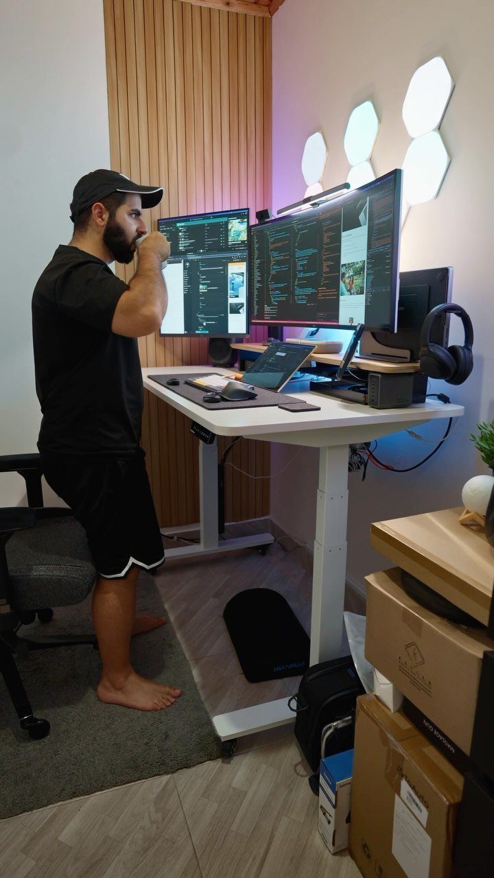 Man standing at a white adjustable desk with dual monitors in a home office.