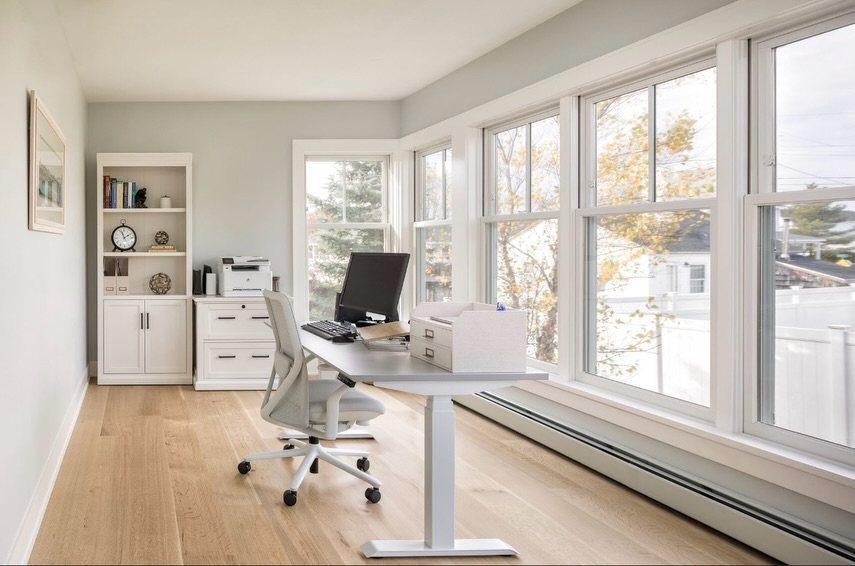 White adjustable desk positioned next to large windows in a bright home office.