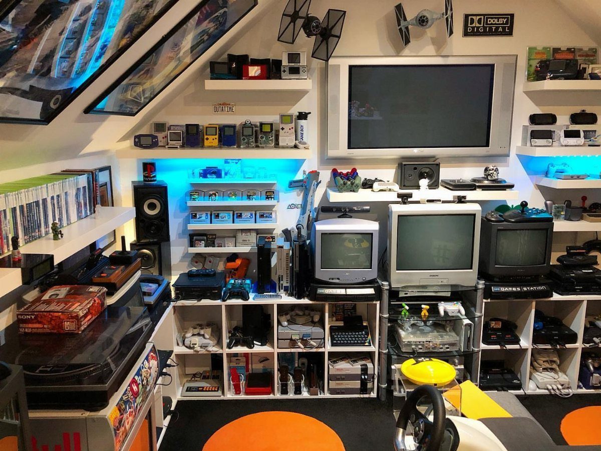 Retro gaming room with CRT TVs, consoles, handhelds, and racing wheel setup.