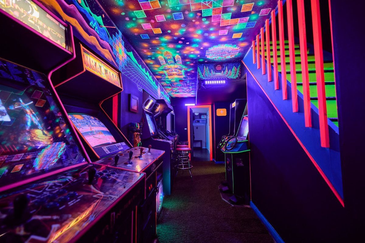 Retro arcade machines in a basement with vibrant neon lighting and colorful ceiling.