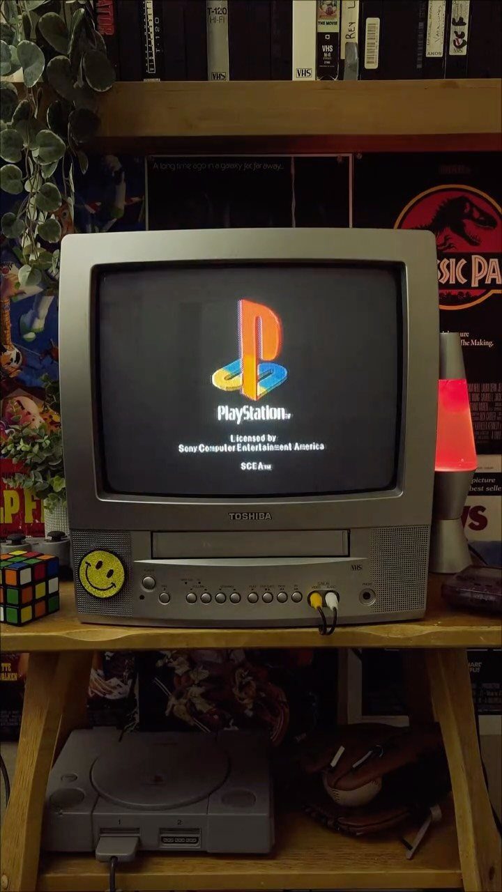 Silver CRT television showing PlayStation logo on a wooden shelf with gaming console.
