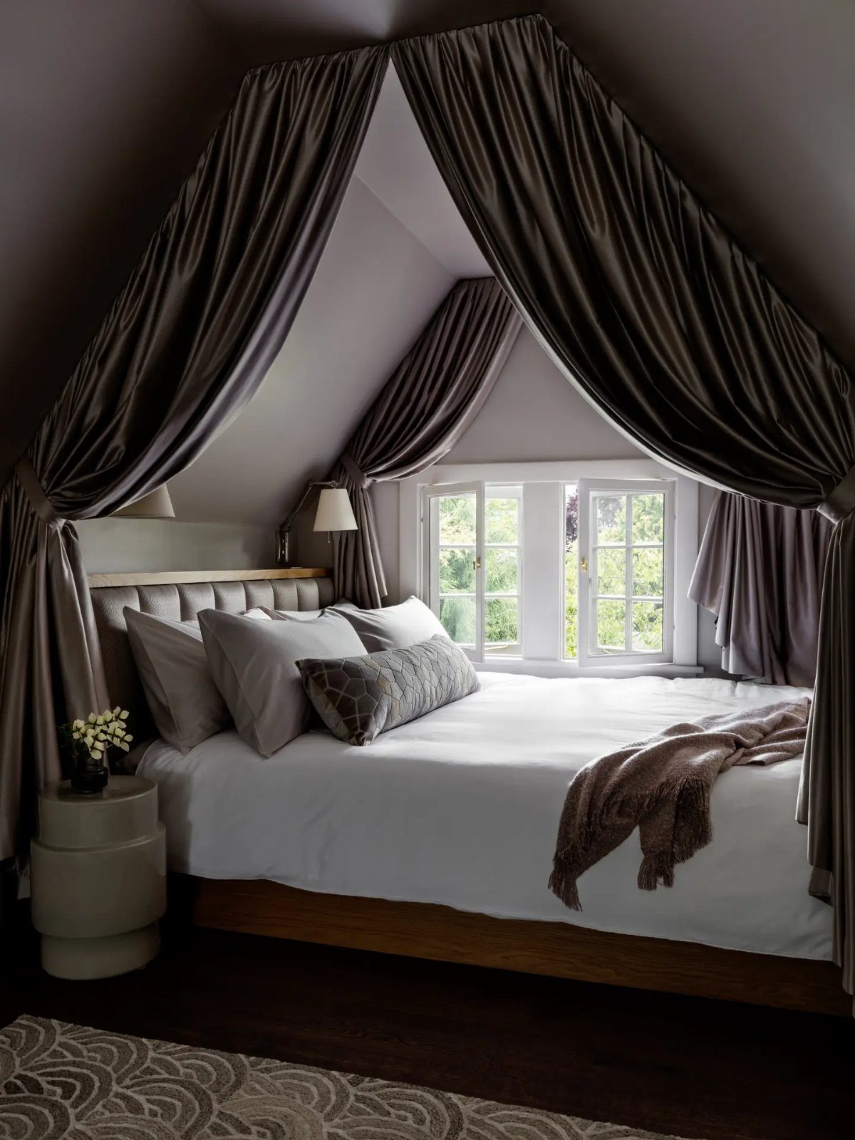 Bed with white linens tucked under a sloped attic ceiling with grey curtains.