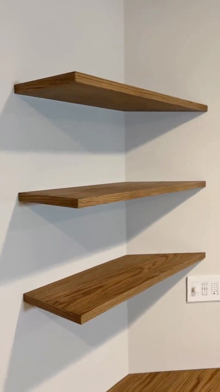 Three light-brown wooden floating shelves mounted on a white corner wall.
