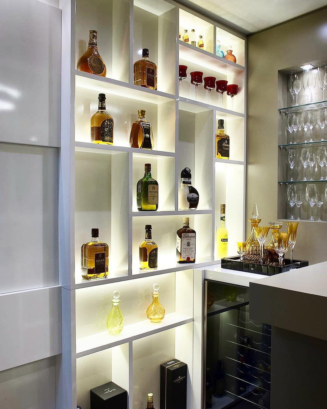 18 Small Bars for Home That Fit Any Space 10 Modern white built-in home bar with backlit asymmetrical shelves and various liquor bottles.
