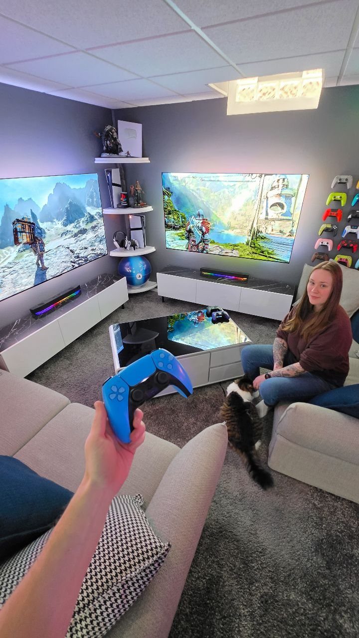 Gaming room with dual screens, white consoles, grey sofa, and a person holding a controller.