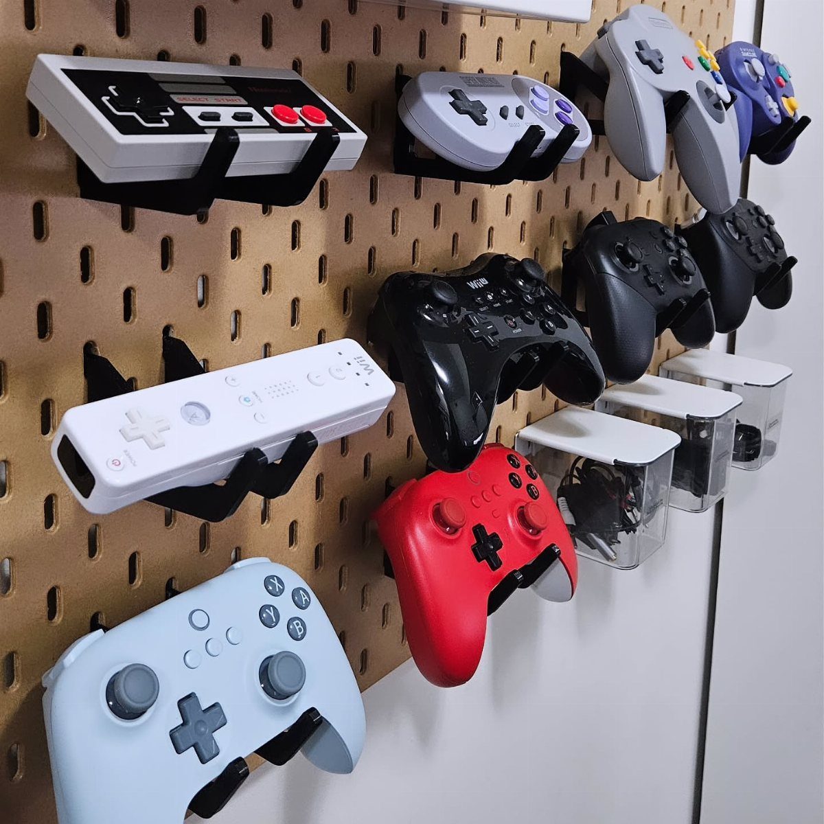 Gaming controllers held by black wall-mounted brackets on a brown pegboard wall.
