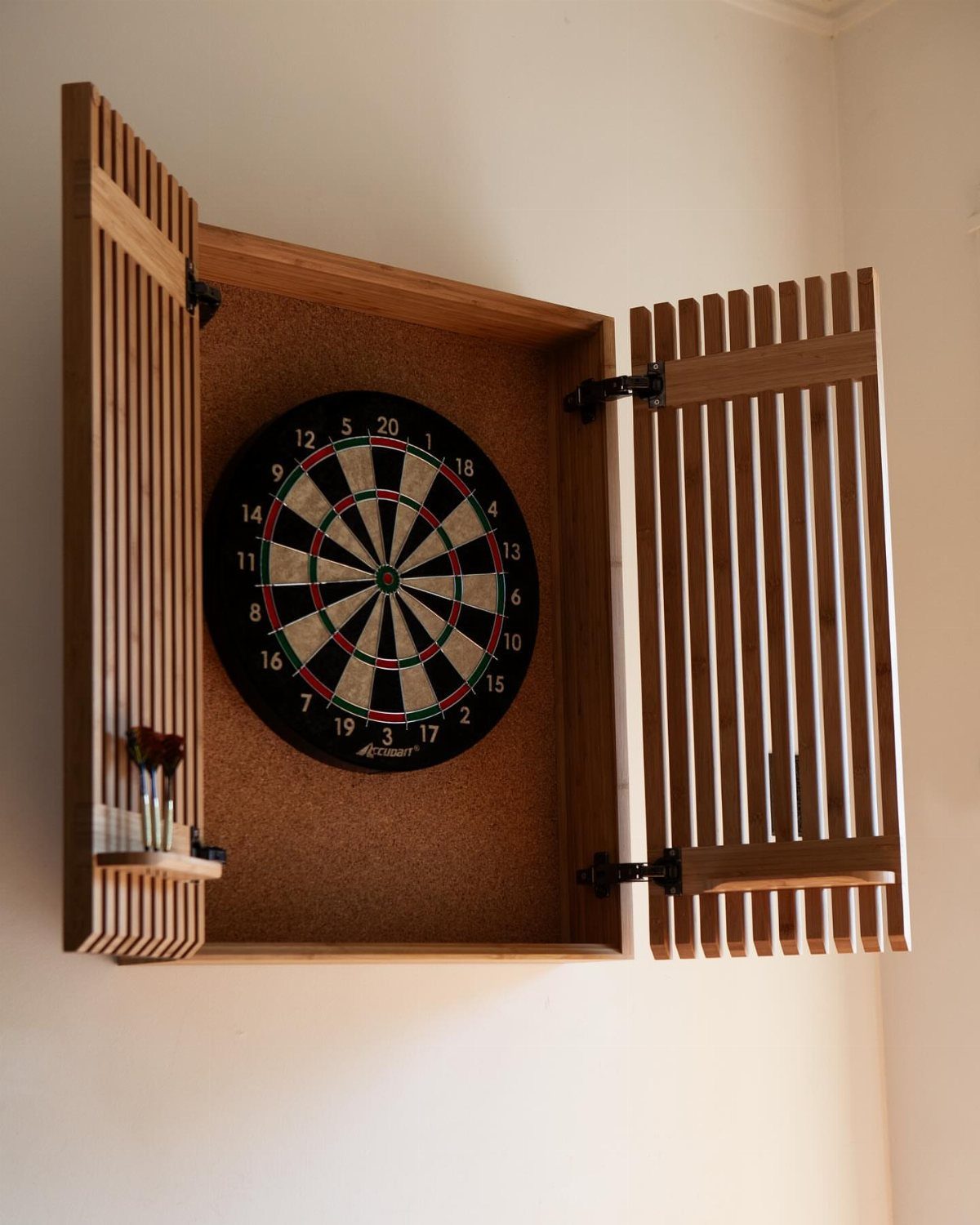 Wooden wall-mounted dartboard cabinet with open slatted doors and a cork backing.