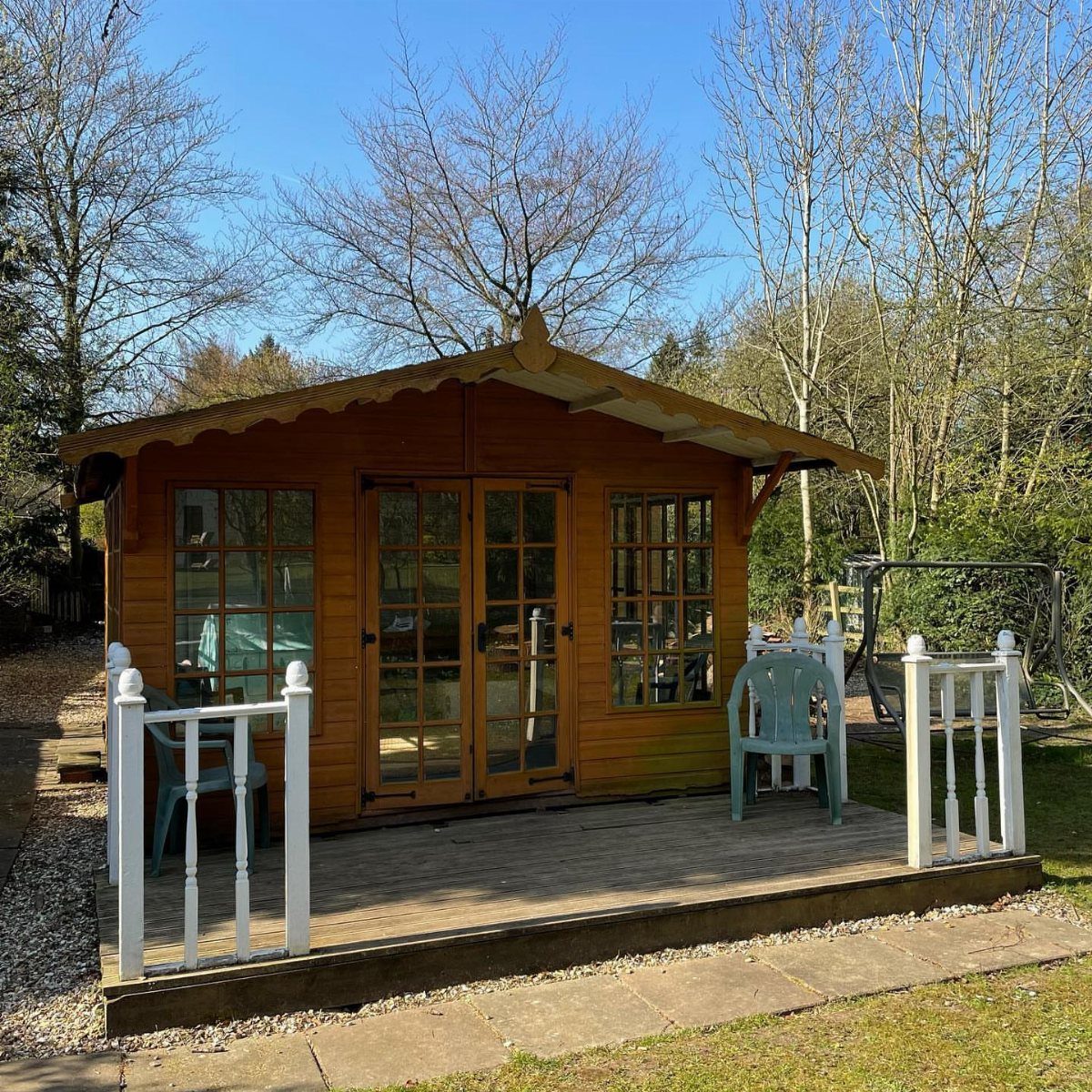 Wooden garden shed with a deck and blue chairs in a sunny backyard