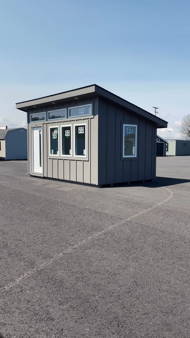 Modern grey shed with white windows and door on an asphalt lot.