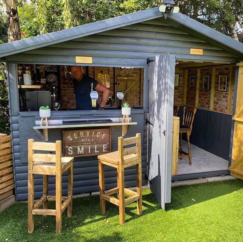 Converted grey garden shed bar with wooden stools and a man serving drinks.