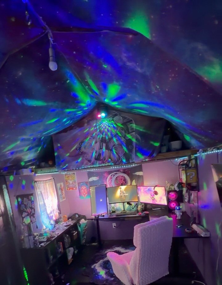 Gaming setup with white chair and monitors under colorful galaxy projection lights.