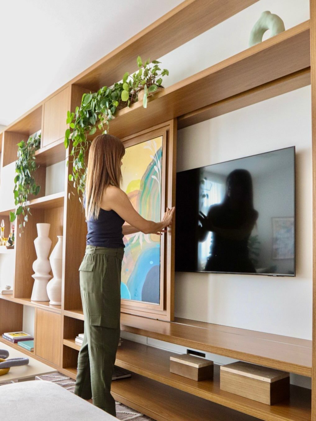 Woman sliding a painting panel to reveal a concealed television in a wooden unit.