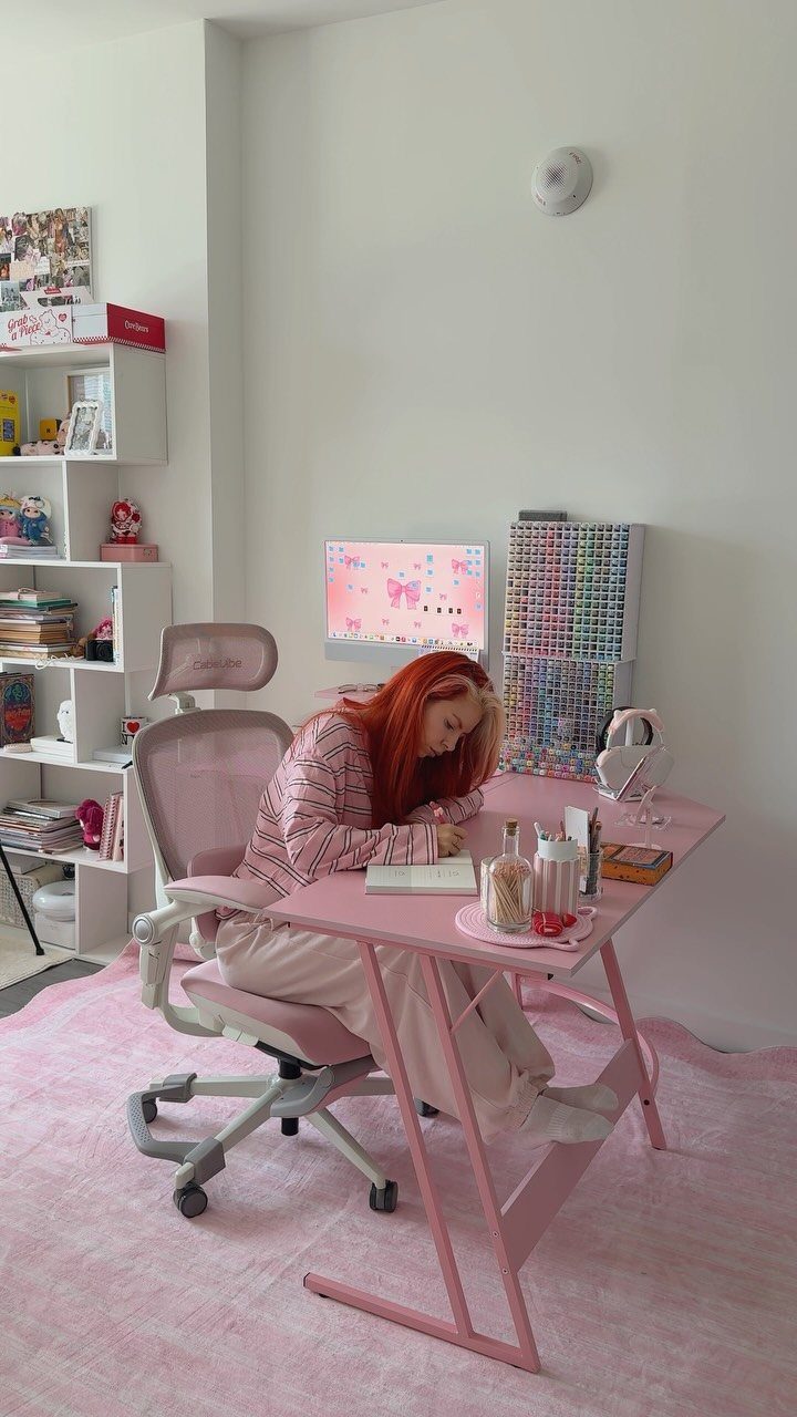 Person with red hair sitting at a pink desk in a bedroom setting.